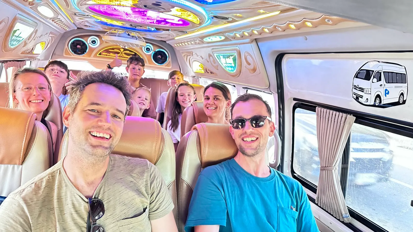 A family group of eight smiling inside Nong’s van on the way to Kanchanaburi